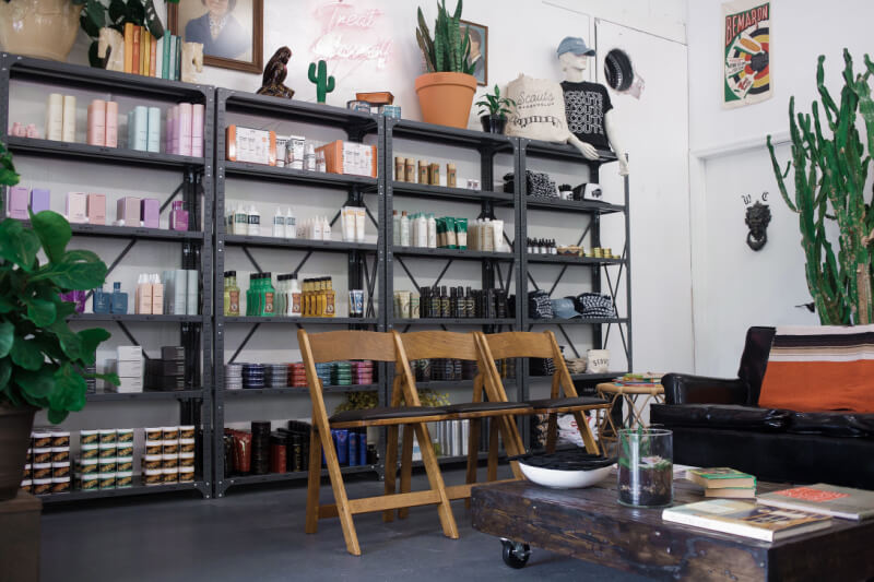 Shelves brimming with beauty products set the scene in this Nashville salon. Two wooden chairs face a sleek black sofa, surrounded by lush plants and chic decor, inviting clients to explore the carefully curated retail selection.