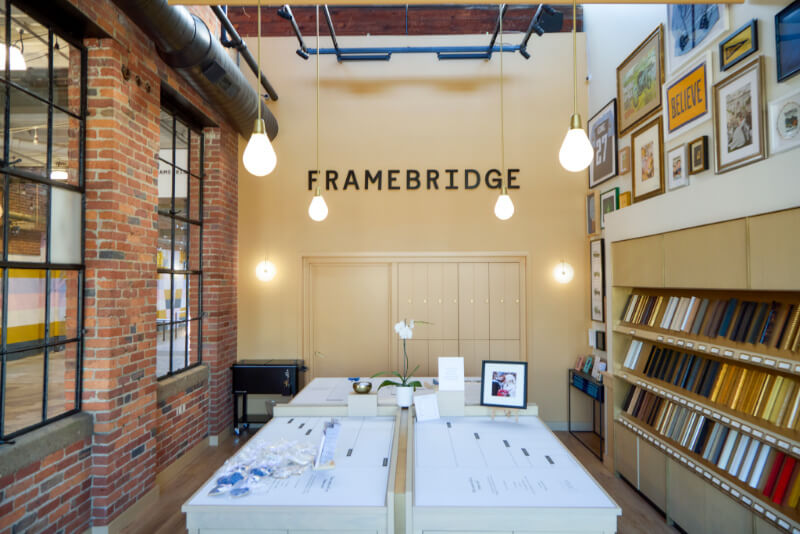 Explore the inviting interior of Framebridge's Nashville store, where brick walls showcase framed artworks. Sample materials are neatly arranged on counters under stylish pendant lights, creating a harmonious retail environment for inspiration and creativity.