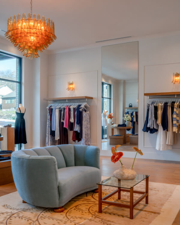 Explore the boutique interior in Nashville featuring a blue sofa, glass coffee table, and hanging clothes. A large mirror and orange chandelier enhance the modern decor, making it a standout retail space.