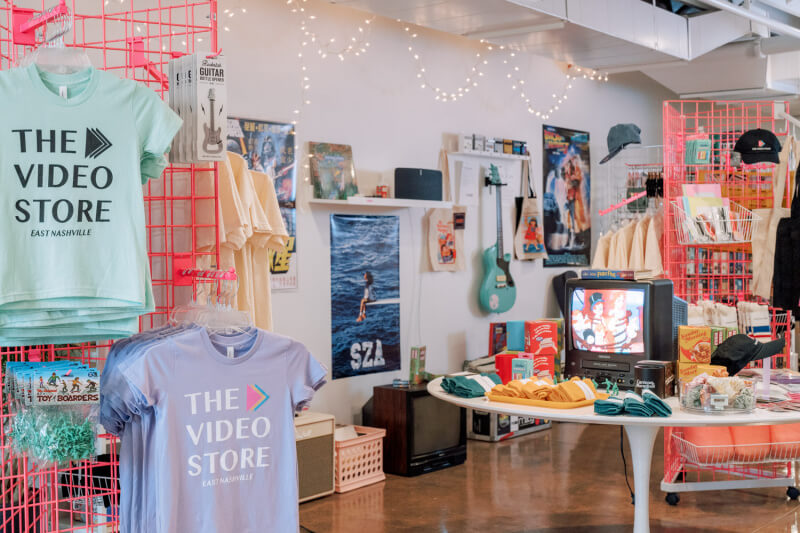 Explore a retro-themed Nashville retail store featuring vintage video cassettes, T-shirts, and memorabilia on wire shelves. A TV playing static sets the vibe, while walls boast posters and a turquoise guitar.
