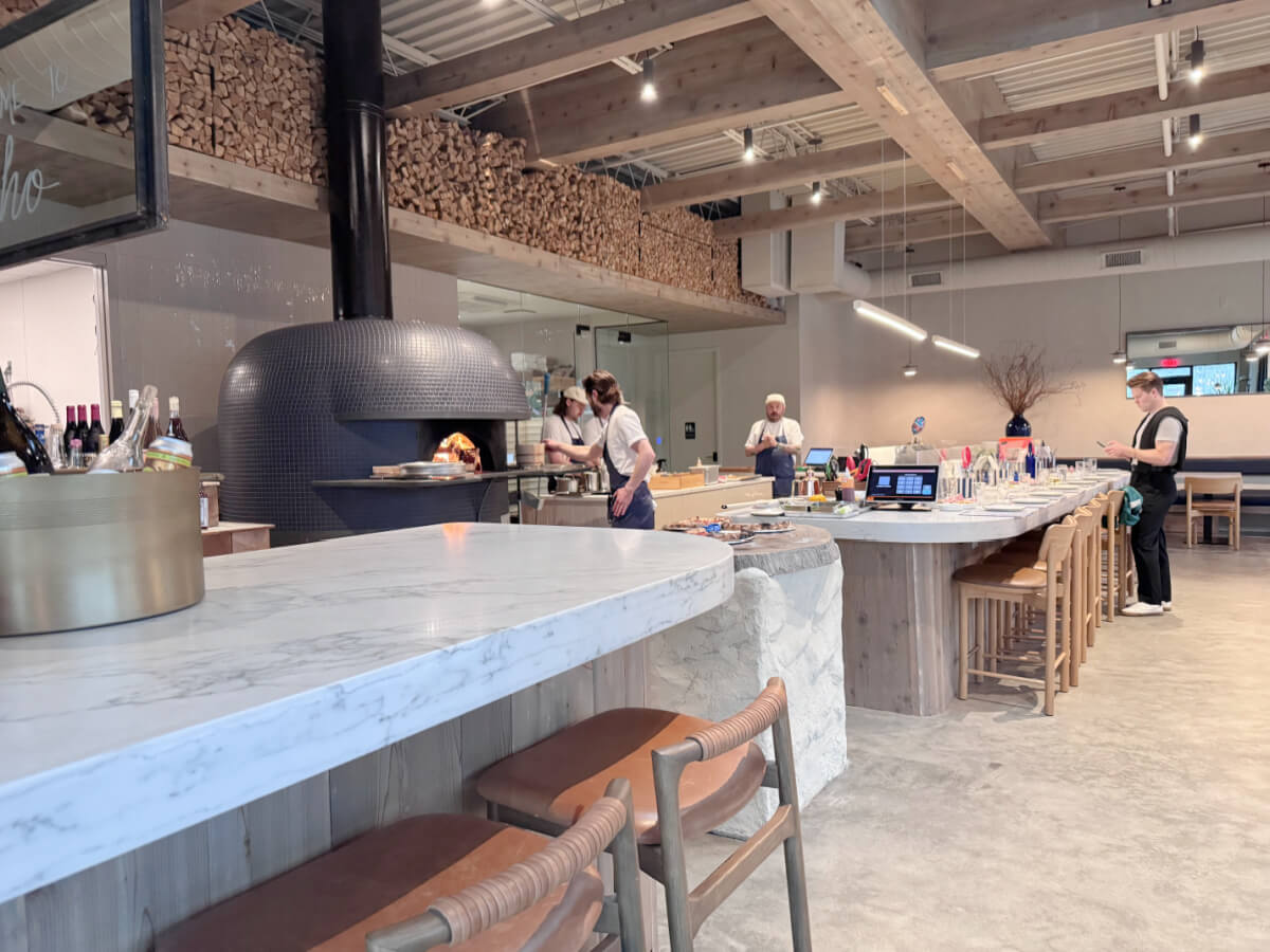 Step into this modern pizza joint, where a wood-fired oven takes center stage alongside bar seating. Watch as the staff crafts delicious creations in an open setting, with stacked wood visible above. 