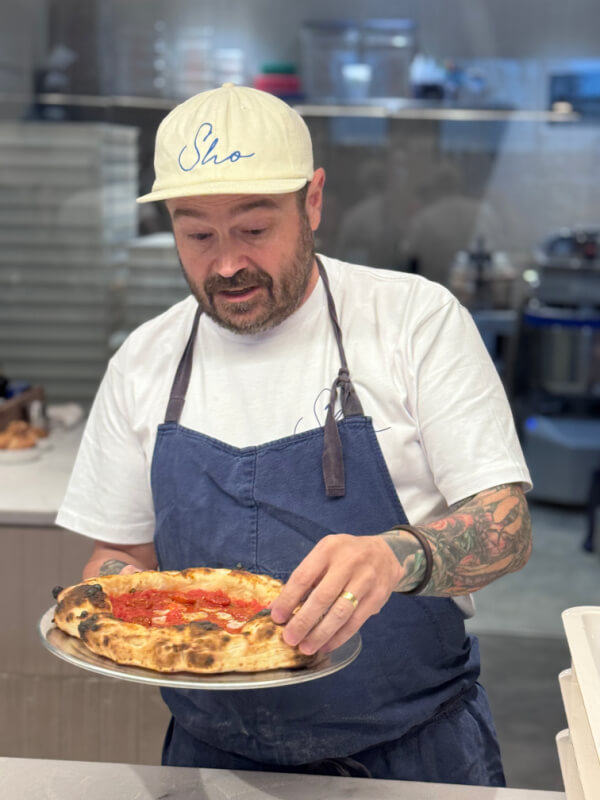 Sean Brock, in a cap and apron, stands proudly at Sho Pizza, holding a freshly made pizza on a metal tray.