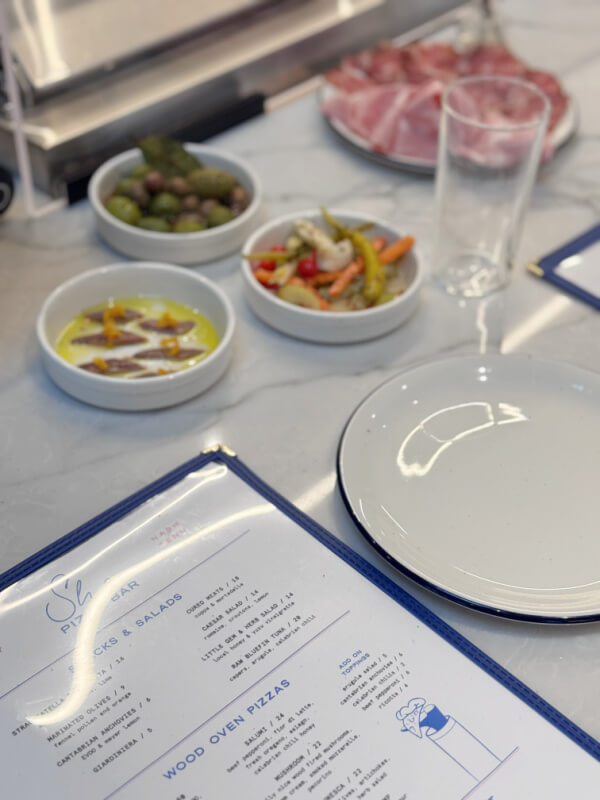 A table set in Sean Brock's restaurant, Sho Pizza, features a menu, an empty white plate, a glass, and small bowls with olives, gardiniera, oil with anchovies, and cold cuts on a marble surface.