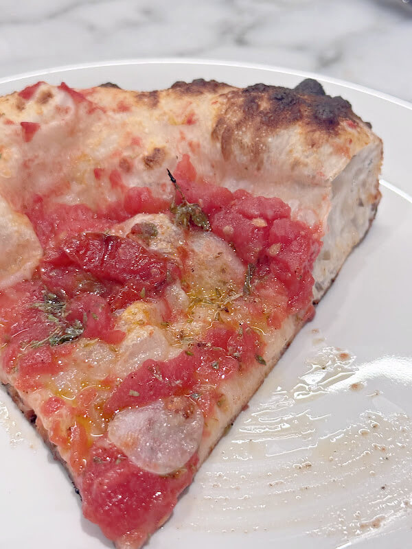 A slice of Sho Pizza's thick-crust delight with tomato sauce, garlic slices, and herbs graces a pristine white plate.