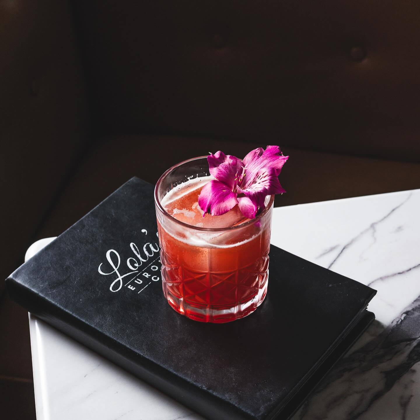 9 New Nashville Restaurants to Try This Month - 24 A red cocktail garnished with a purple flower sits on a black menu atop a marble table.