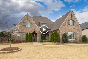 A large brick house in Collierville, TN, with a steep roof, manicured shrubs, and a tree on the front lawn under a cloudy sky.