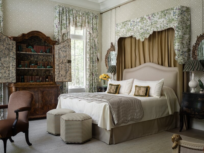 A traditional bedroom showcased in the 2025 Southeastern Designer Showhouse features a canopy bed, two striped ottomans, floral curtains, an open antique cabinet with books, and a window letting in natural light.