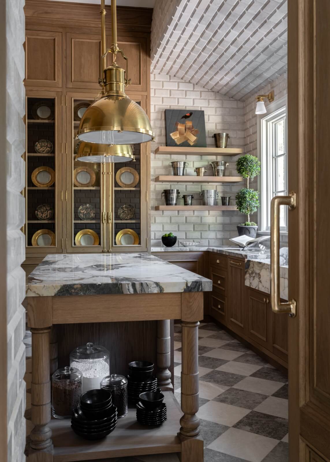 A stylish kitchen features wood cabinets, marble countertops, brass light fixtures, open shelves with plants, and black dishes on a lower shelf. Walls and ceiling are clad in white brick.