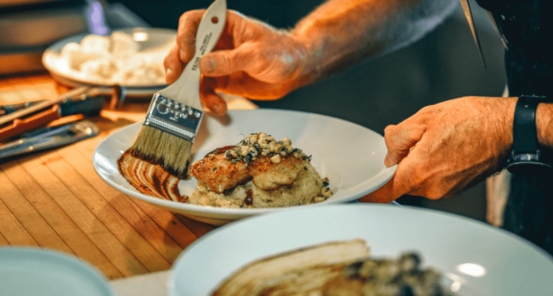 In a kitchen setting, someone applies sauce with a brush to a plated dish of chicken on risotto, delicately garnished with herbs. 