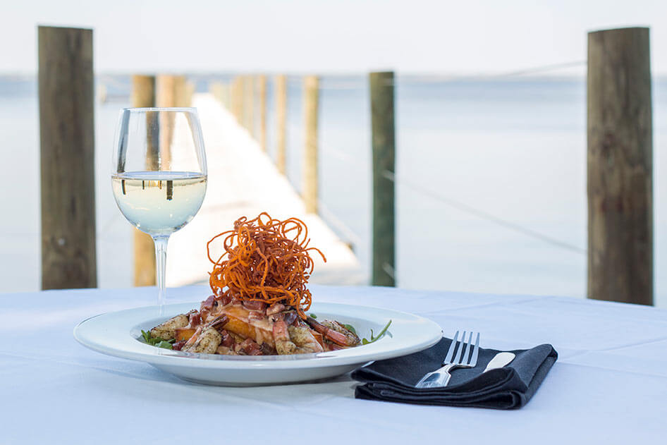 A plate of shrimp with crispy garnish and a glass of white wine sit elegantly on a table overlooking the dock, encapsulating the charm of 30A dining. 