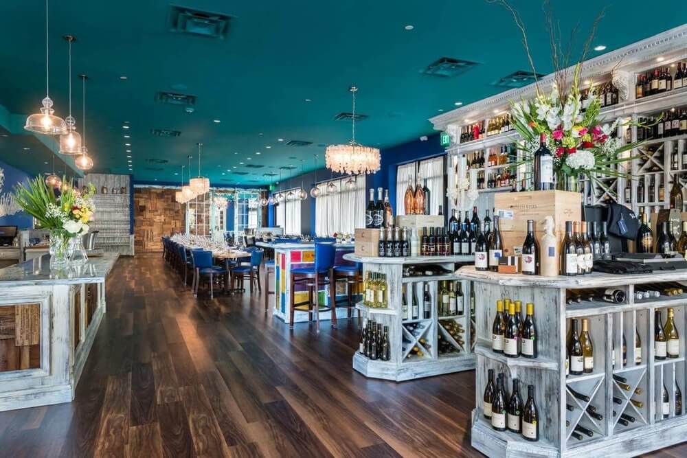 Step into the elevated dining experience of this contemporary restaurant. With wooden flooring, tables elegantly set for dining, and shelves of fine wine bottles, all under a teal ceiling adorned with chandeliers and floral arrangements.