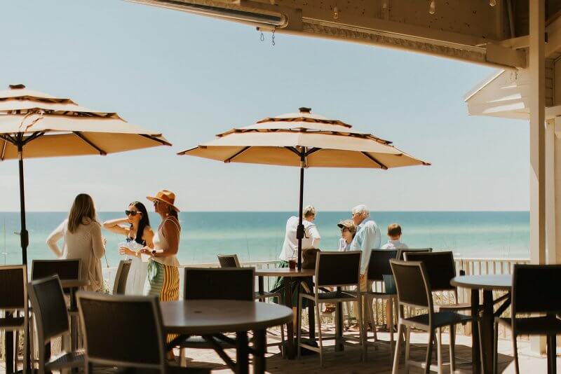 People enjoy a sunny day at a beachside café on 30A, under large umbrellas, overlooking the ocean. Consider booking ahead for an elevated dining experience that perfectly complements the serene seaside view.