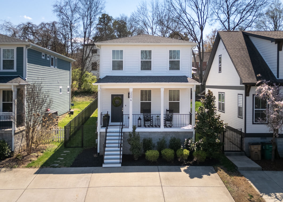 A charming two-story white house at 2509A Solon Dr, Nashville, TN, features a welcoming front porch with black chairs. It boasts a neatly landscaped yard and is surrounded by similar style homes, complete with a paved driveway in front.