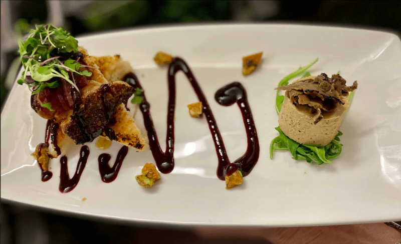 Gourmet dish with pâté on greens, toast with garnish, and drizzled sauce presents an elevated dining experience on a white rectangular plate.