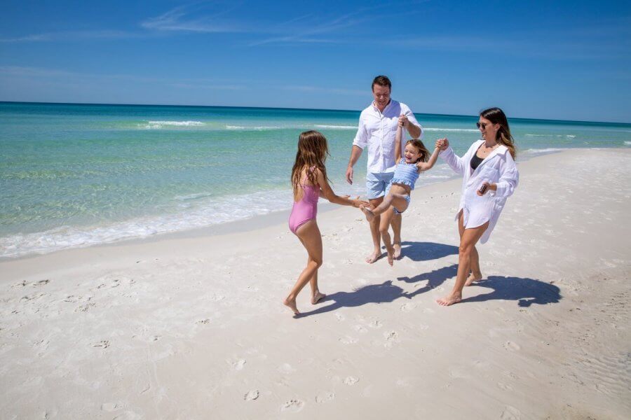 5 Reasons South Walton is Becoming the Premier Destination for Corporate Events and Meetings - 6 A family of four enjoys an event in South Walton, walking on a sandy beach by the ocean; two adults swing a young child while another walks nearby under the clear blue sky.