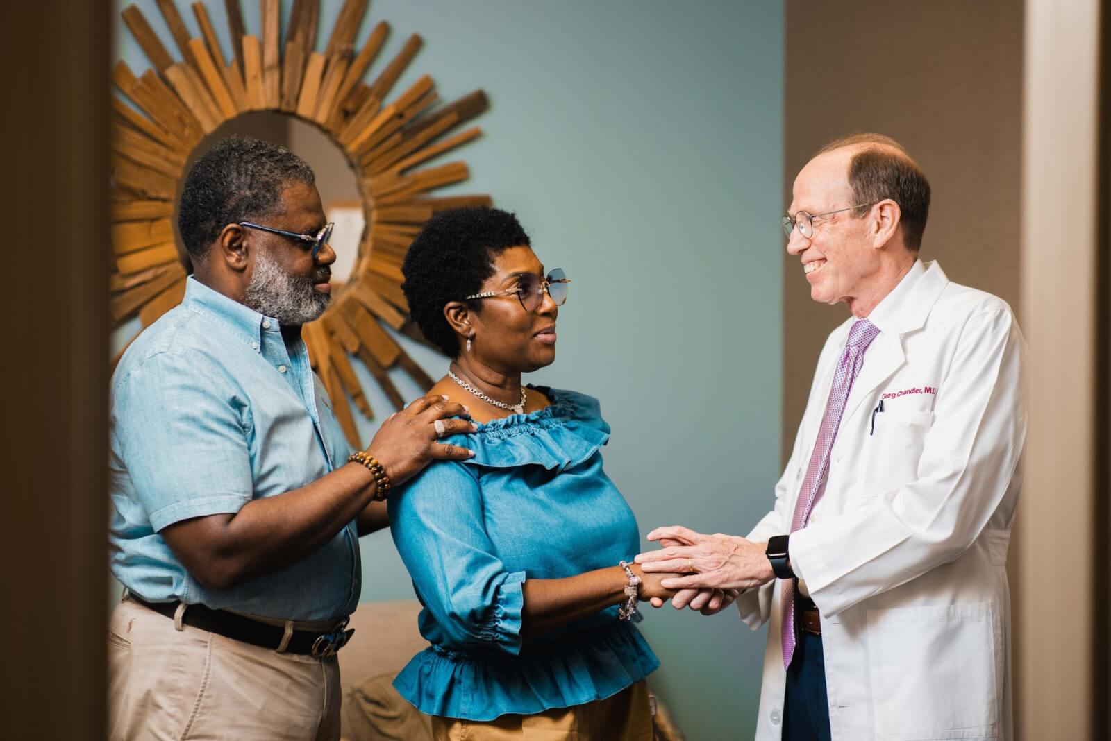 A doctor in a white coat speaks with a woman and a man at Plastic Surgery Group Memphis. The man stands beside the woman with his hand on her shoulder. A decorative sunburst mirror is on the wall behind them.