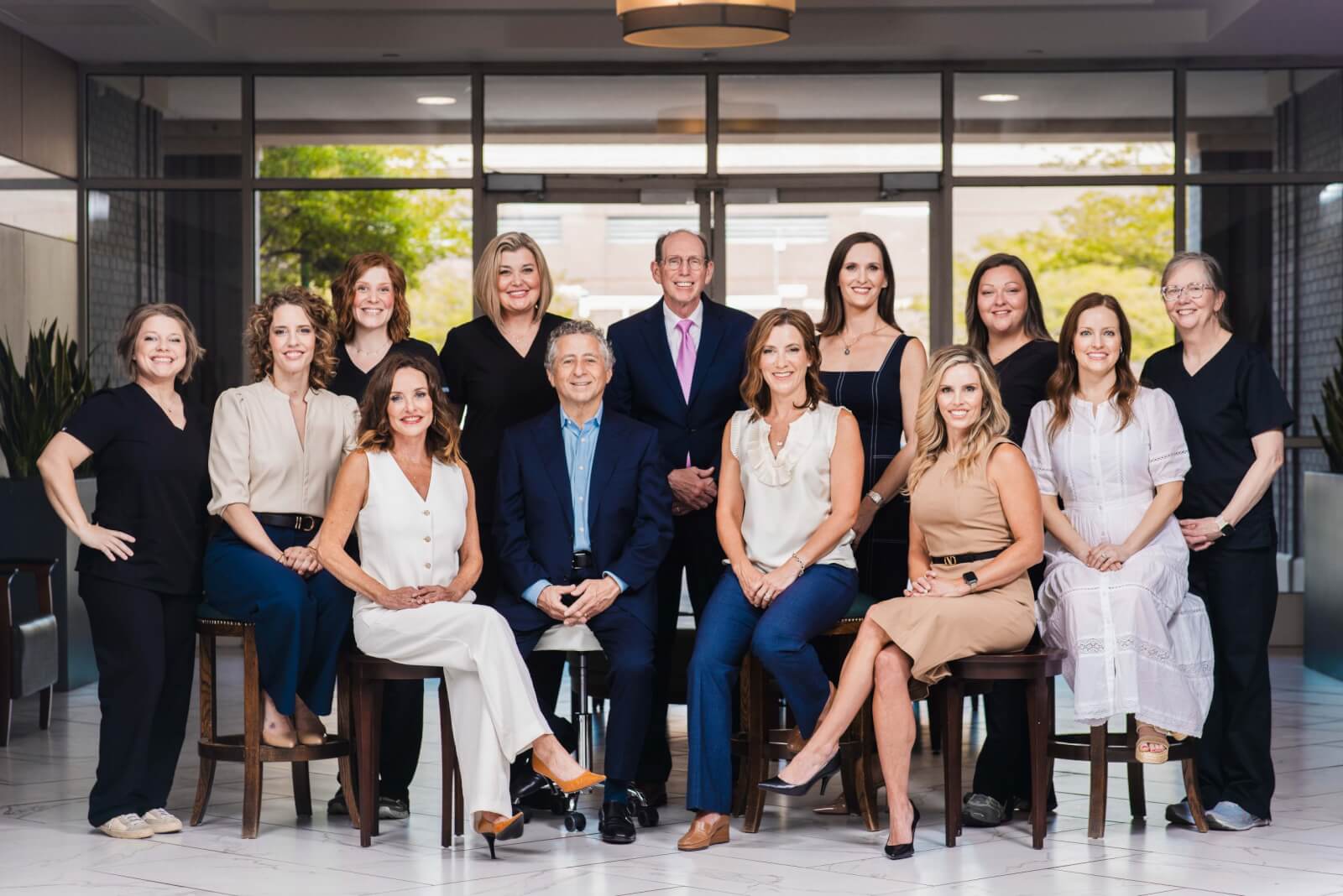 A group of thirteen adults, dressed in business and casual attire, pose together indoors in two seated and standing rows, with large windows behind them at a Plastic Surgery Group in Memphis.