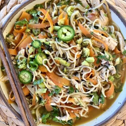 A High Protein Noodle Bowl with shredded chicken, sliced carrots, green onions, cilantro, and jalapeño slices, served with chopsticks on a woven placemat—perfect for a quick dinner.
