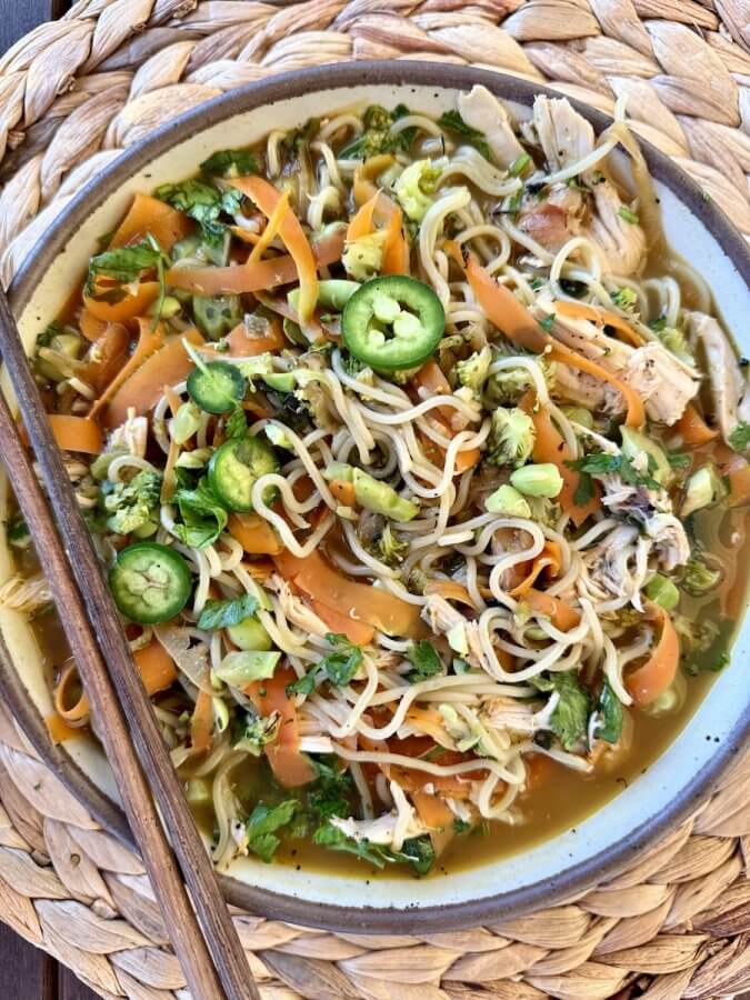 A High Protein Noodle Bowl with shredded chicken, sliced carrots, green onions, cilantro, and jalapeño slices, served with chopsticks on a woven placemat—perfect for a quick dinner.