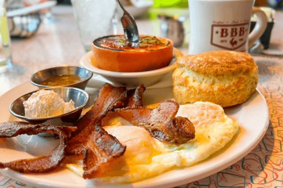 A Big Bad Breakfast plate with two sunny-side-up eggs, bacon, a biscuit, jelly, butter, coffee, and a baked tomato topped with herbs.