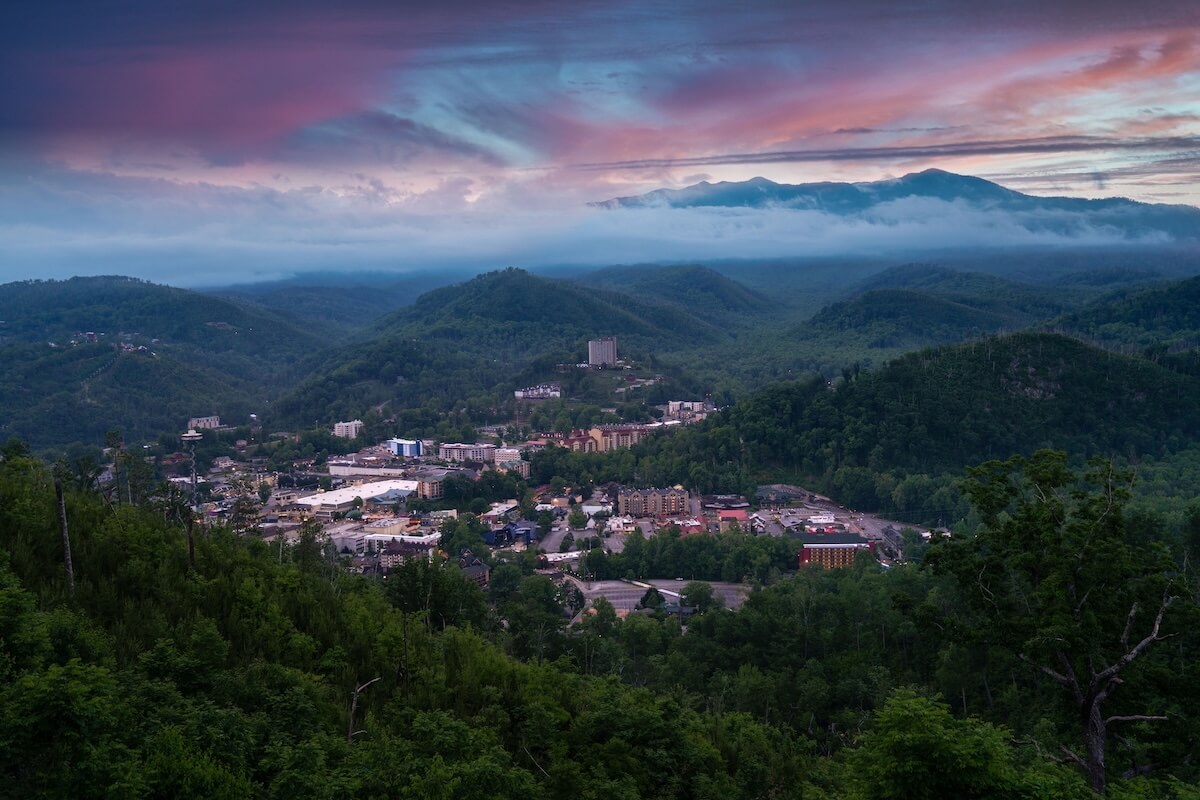 Scenic mountain town in TN with buildings nestled among green hills under a colorful, cloudy sunset sky, mist curling around the distant mountains—an inviting Gatlinburg getaway.
