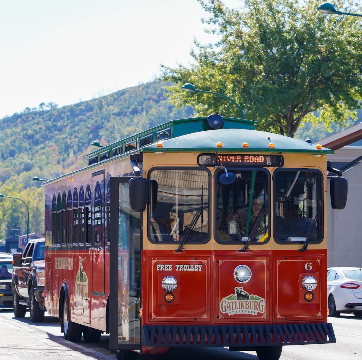 A red Gatlinburg trolley labeled "River Road" is stopped on a street in TN, with trees and mountains in the background—perfect for your next getaway.