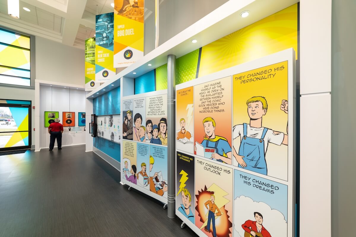 A museum or exhibit space features large comic-style panels on the walls, depicting personal change narratives with bright illustrations and captions—perfect inspiration for a girls' trip or travel planning adventure in Tupelo MS. A person views a display in the background.