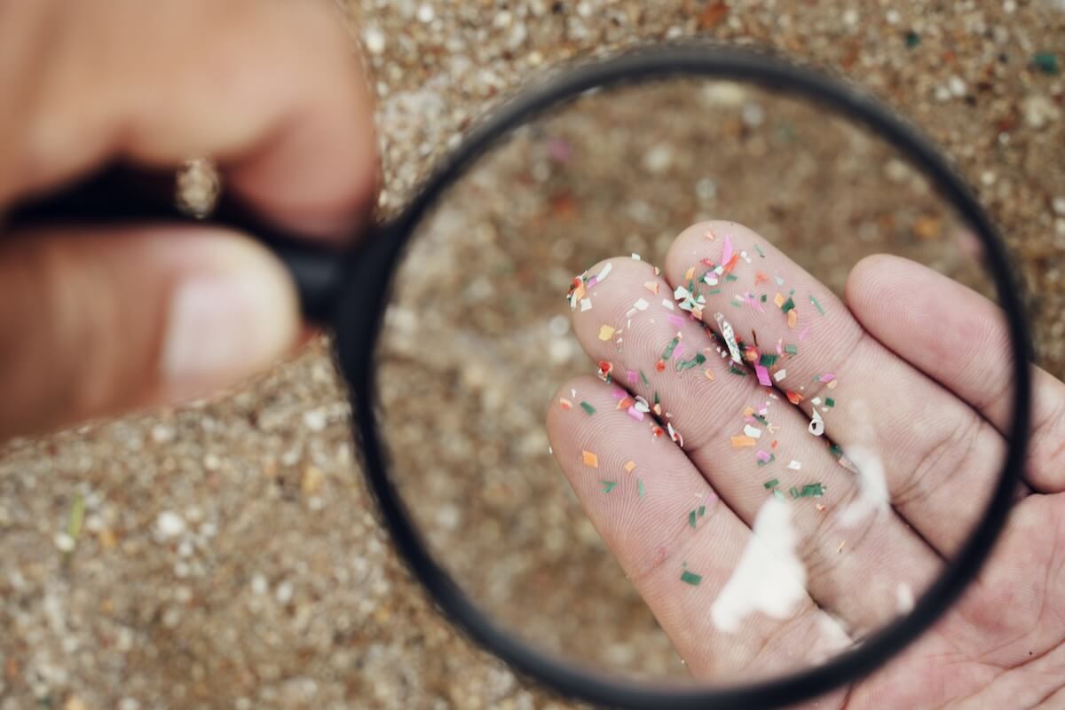 Shocking Stats About Microplastics You Need to Know (Seriously.) - 1 A hand holding small colorful microplastics is viewed through a magnifying glass against a sandy background, highlighting their environmental impact.