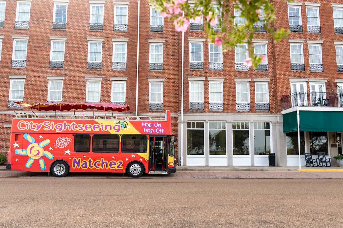 Why Natchez, MS, Belongs on Your Summer Travel List - 5 A red City Sightseeing Natchez tour bus is parked in front of a brick building with large windows and an awning on an empty street in Natchez, MS.