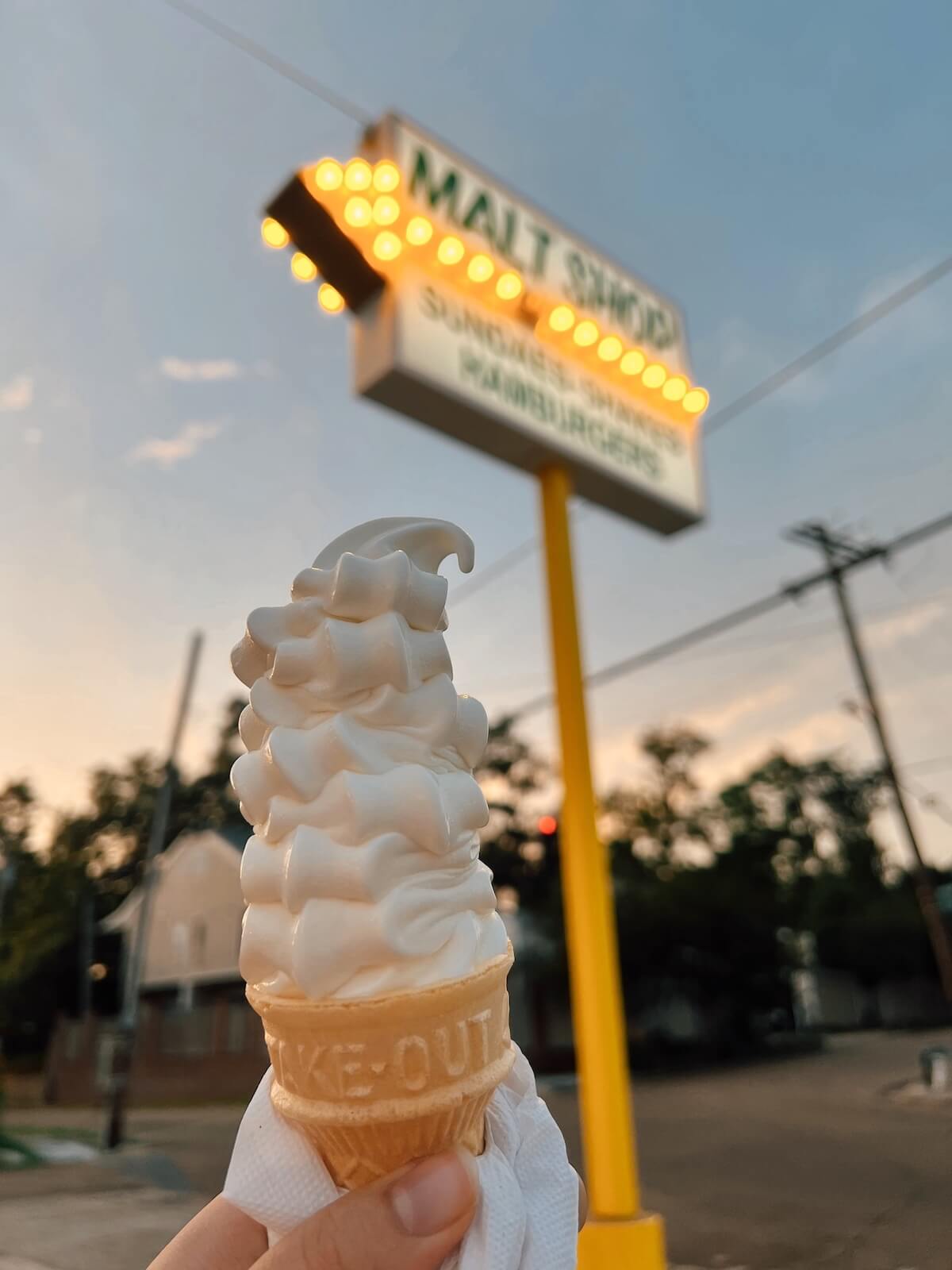 Why Natchez, MS, Belongs on Your Summer Travel List - 4 A hand holds a soft-serve ice cream cone in front of a blurred malt shop sign at dusk in Natchez MS, with trees and power lines behind—a perfect summer travel moment for your travel list.