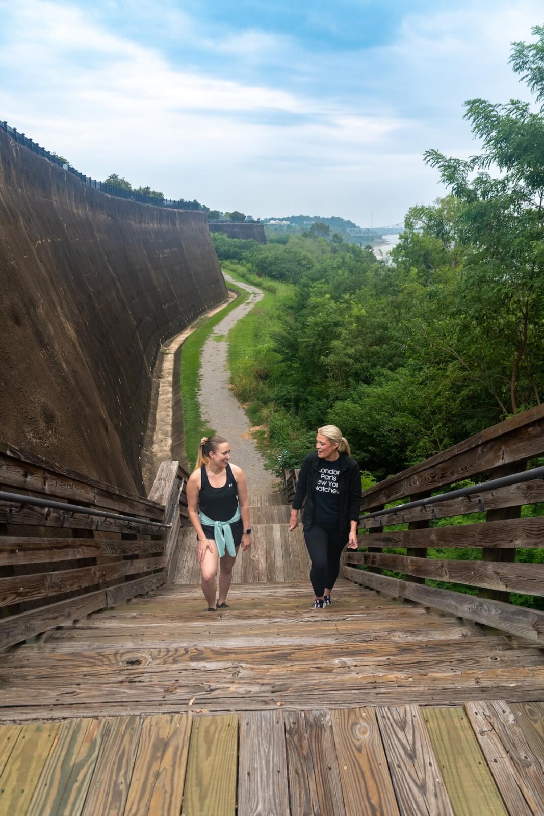 Why Natchez, MS, Belongs on Your Summer Travel List - 2 Two women walk up wooden stairs next to a large concrete wall in Natchez MS, with greenery and a path behind them under a partly cloudy sky—a perfect spot for your summer travel list.