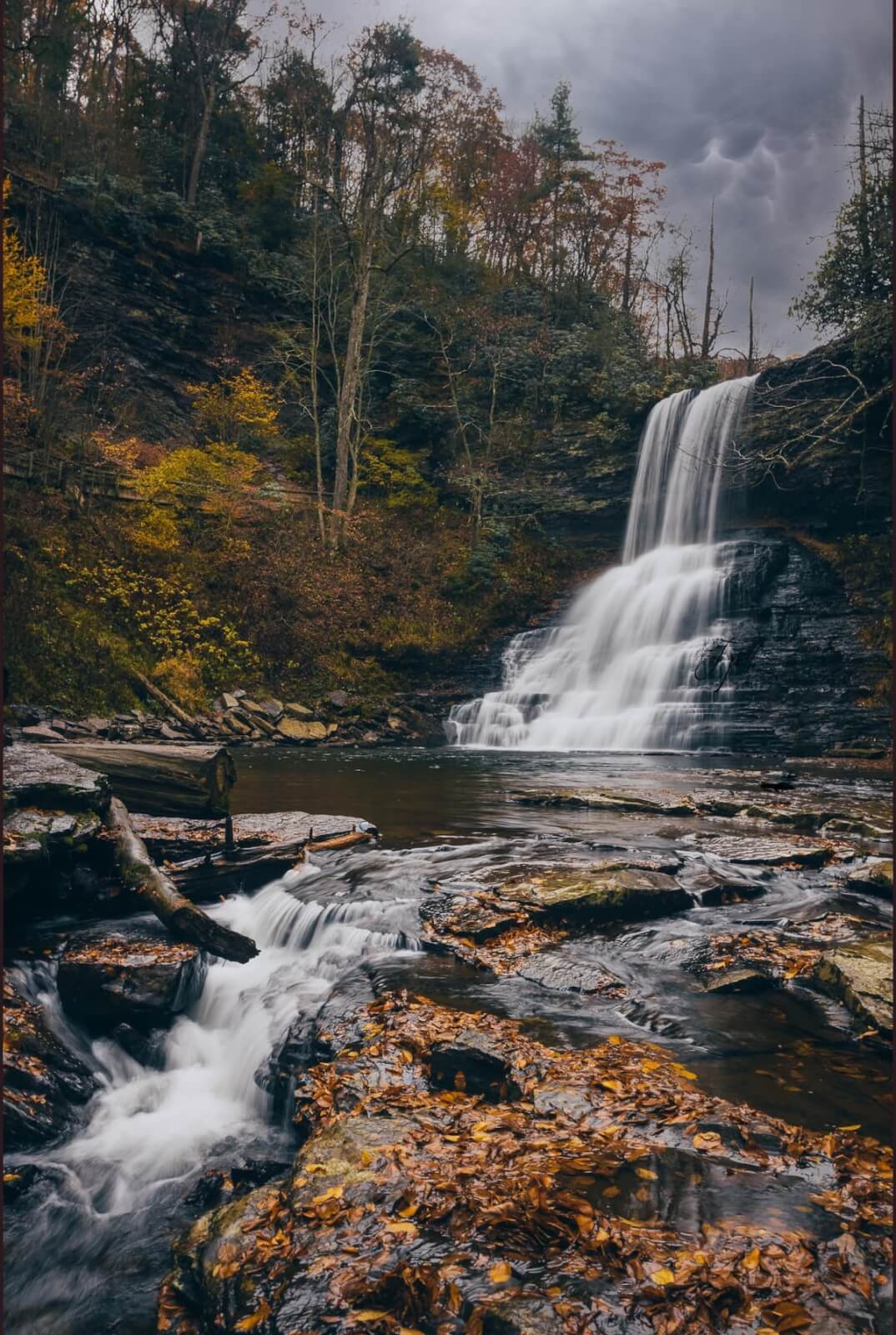 A waterfall cascades over a rocky cliff into a stream surrounded by autumn trees and fallen leaves under a cloudy sky—perfect for scenic waterfall hikes in Virginia.