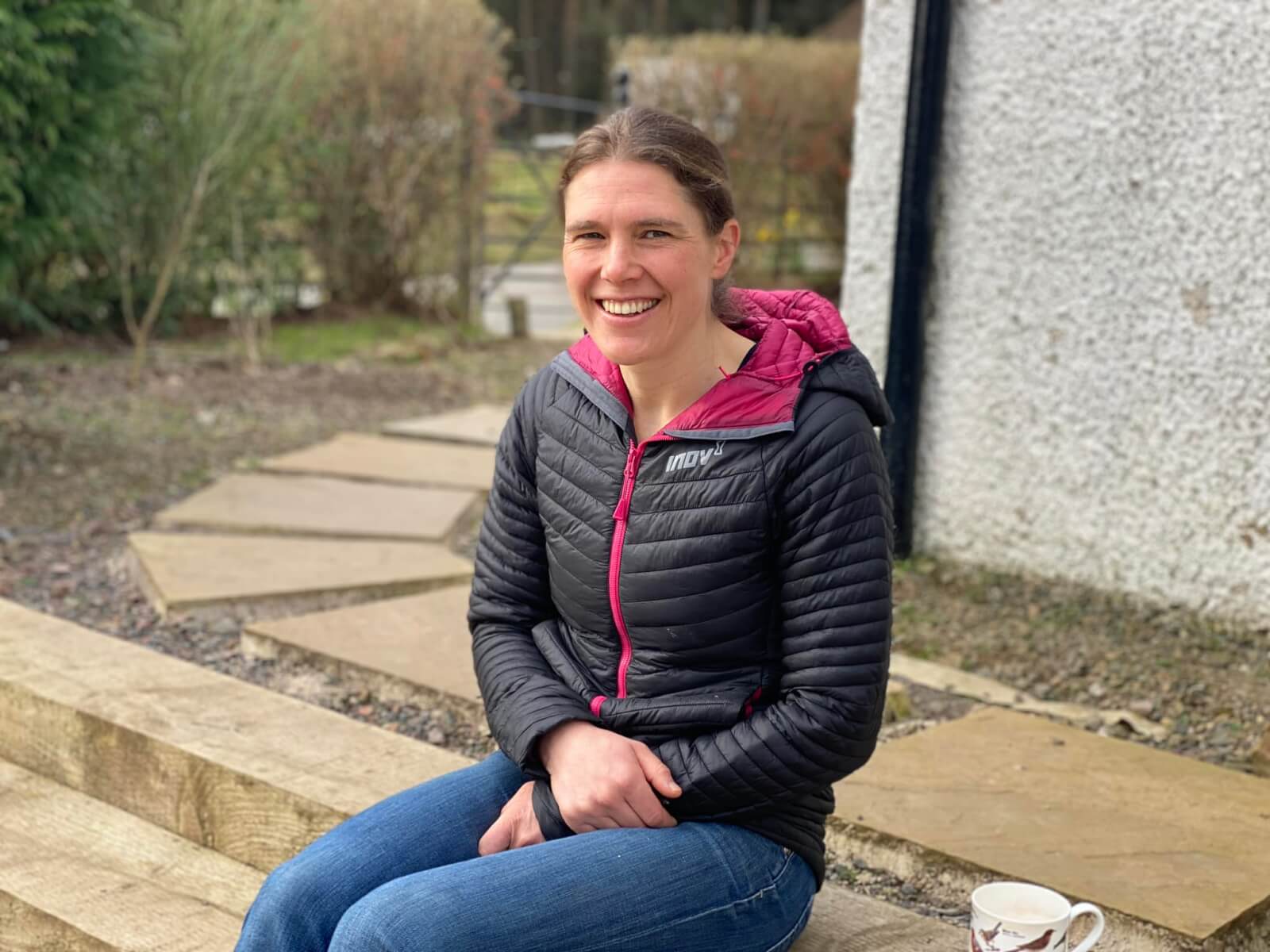 A woman wearing a black puffer jacket and jeans sits on outdoor steps at Frozen Head State Park, smiling at the camera. A mug is placed on the ground nearby, evoking the spirit of the Barkley Marathons.
