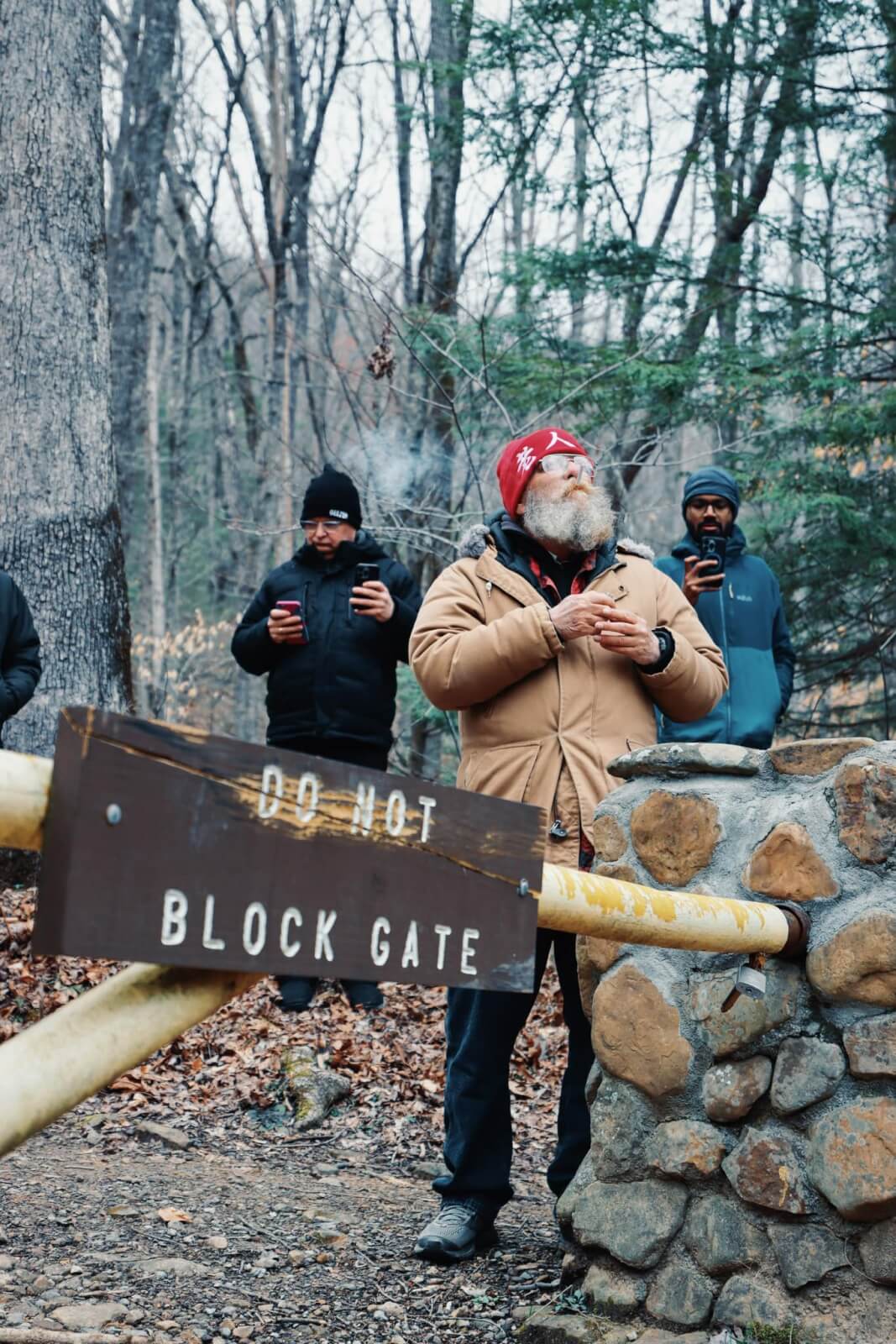 Four men stand near a stone gate in a wooded area at Frozen Head State Park, with one man in a tan jacket and red hat in front. A sign reads "DO NOT BLOCK GATE," hinting at the legendary Barkley Marathons held here.