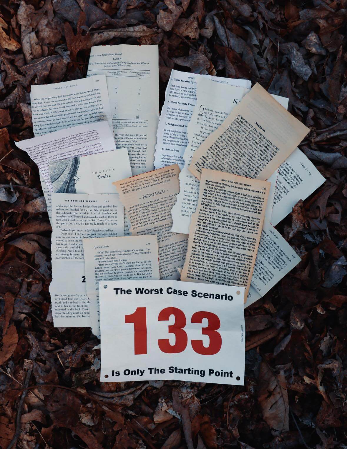 A stack of torn book pages lies on dry leaves at Frozen Head State Park, with a race bib labeled "133" and the words "The Worst Case Scenario Is Only The Starting Point," evoking the spirit of the Barkley Marathons.