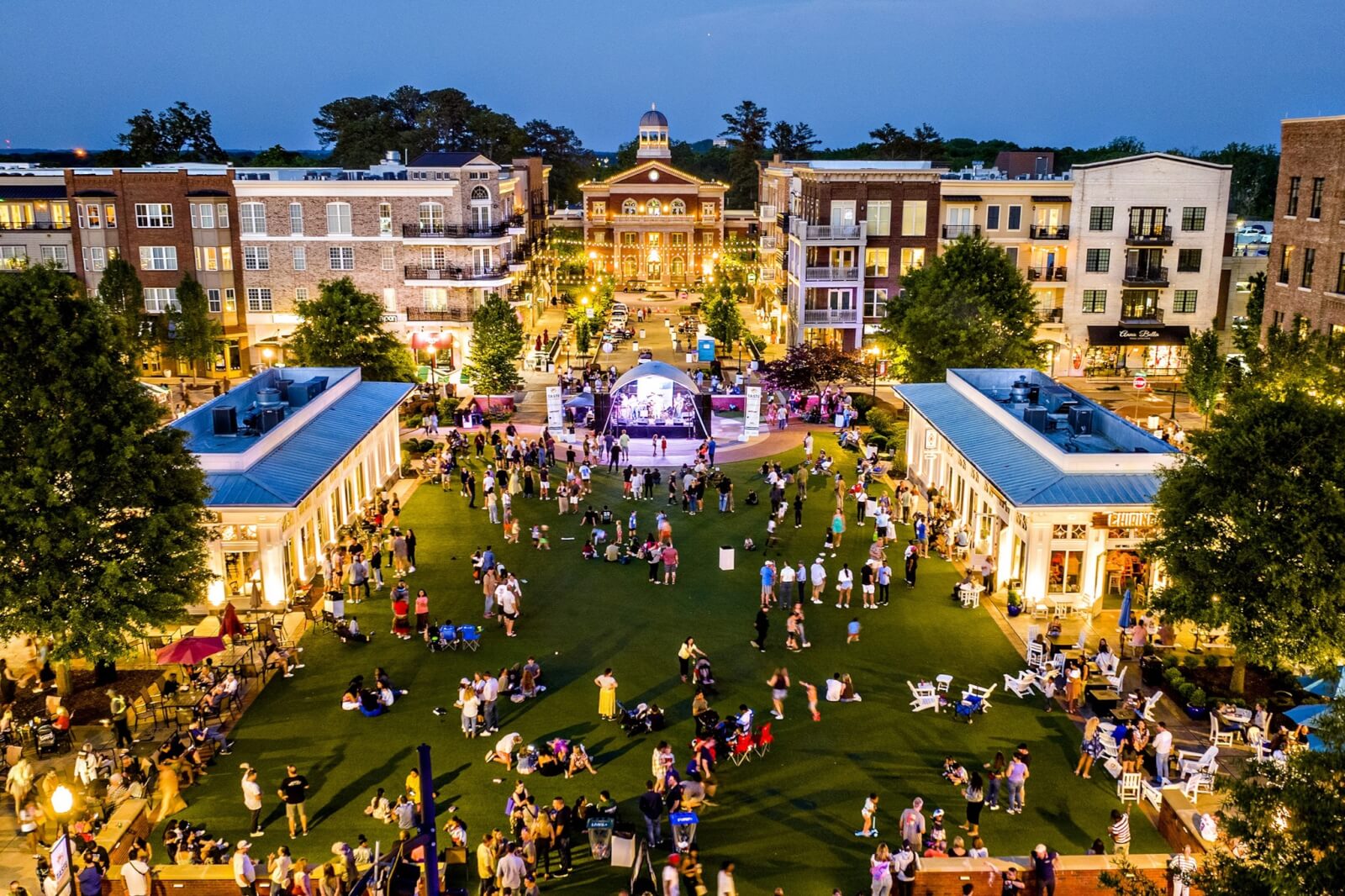 Georgia's 6 Best Girlfriends' Weekend Getaways - 3 Aerial view of a lively outdoor gathering at a town square with people, grass lawn, stage, shops, and multi-story buildings at dusk—perfect for girlfriends getaways or relaxing Georgia getaways.