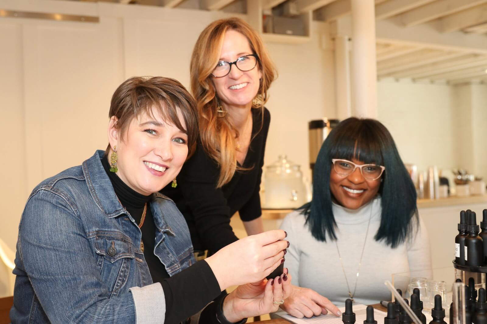 Georgia's 6 Best Girlfriends' Weekend Getaways - 1 Three women sit and stand around a table with bottles and droppers, smiling at the camera in a bright, modern room—capturing the joy of friendship during weekend getaways in GA.