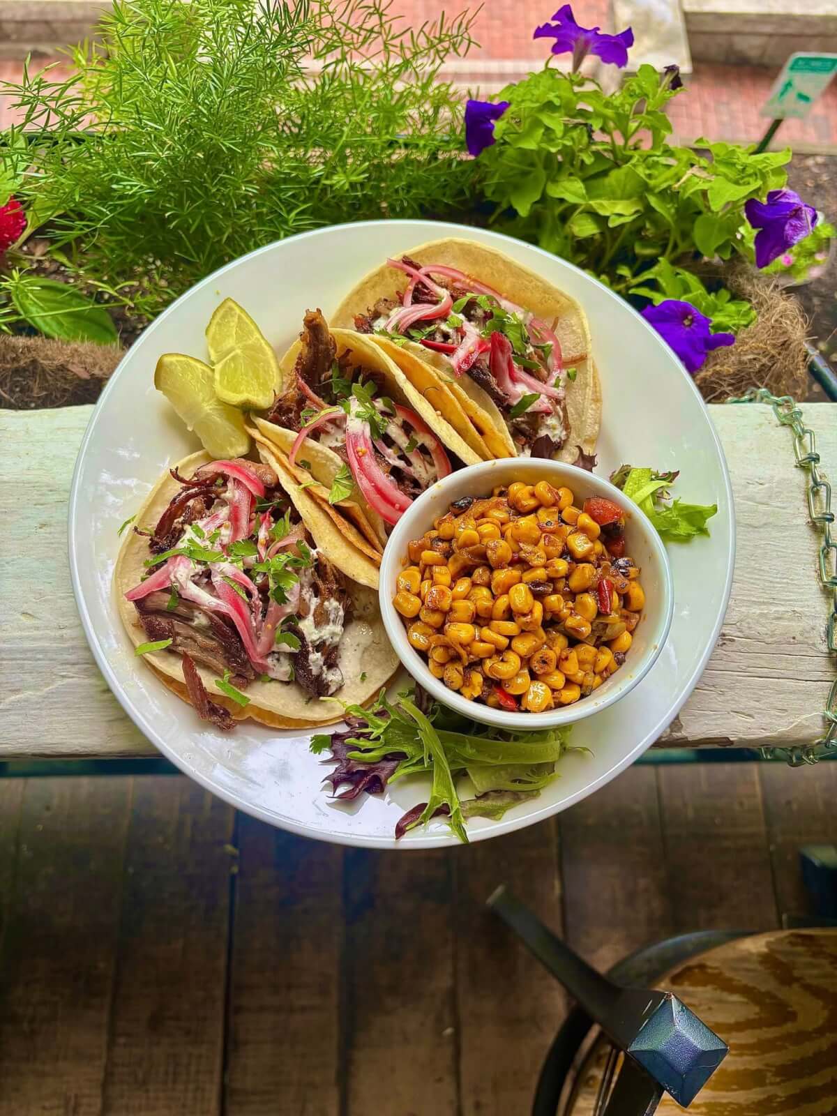 Georgia's 6 Best Girlfriends' Weekend Getaways - 9 A plate with three tacos topped with shredded meat, pink onions, and cilantro, served with lime wedges, roasted corn, and a small salad, set outdoors near lush plants.