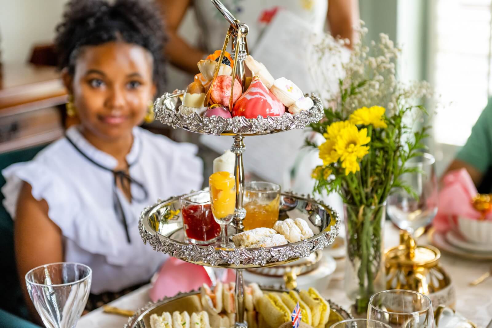 Georgia's 6 Best Girlfriends' Weekend Getaways - 2 A tiered tray of assorted pastries and sandwiches sits on a table with flowers, glasses, and tea—perfect for relaxing moments during weekend getaways in GA, with a woman seated in the background.