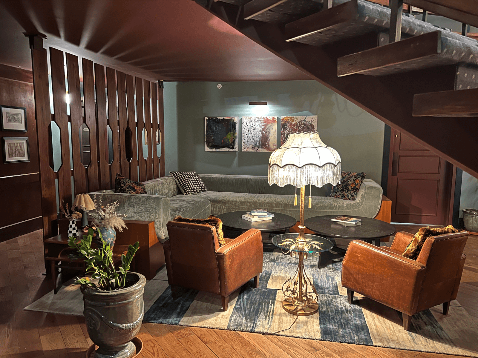A cozy living room in this boutique Chattanooga hotel features a sectional sofa, two brown armchairs, a fringed floor lamp, round tables, plants, and framed art—inviting you to unwind in style on your solocation, as seen from under the staircase.