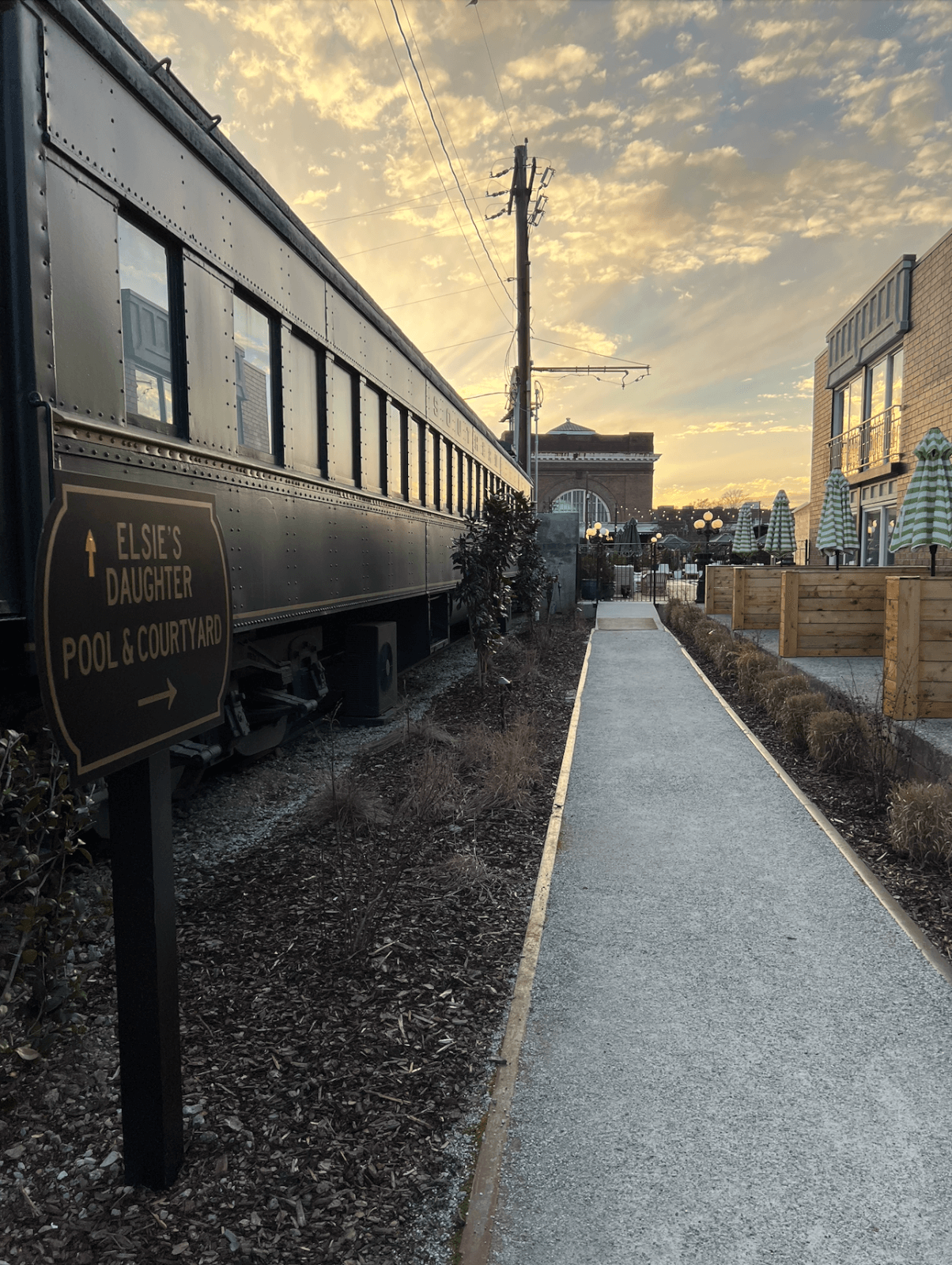 A paved path runs alongside a vintage train car at this boutique Chattanooga hotel, with a sign pointing to "Elsie's Daughter Pool & Courtyard"; outdoor seating and buildings are visible under a sunset sky.