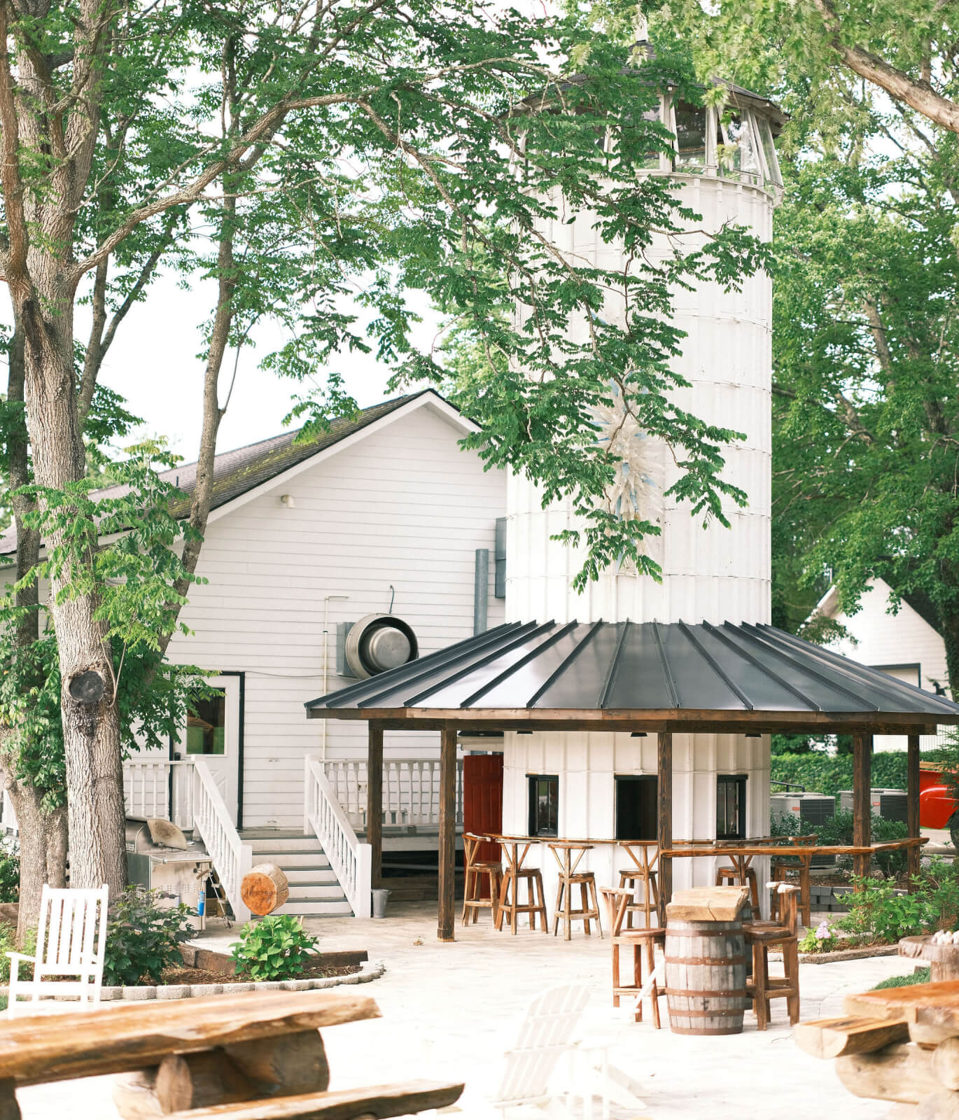 Charming white farmhouse-style building with a tall, silo-like structure, nestled among trees in Hendersonville NC. Experience summer relaxation with outdoor seating—wooden benches, tables, and barrels—at this boutique farm.