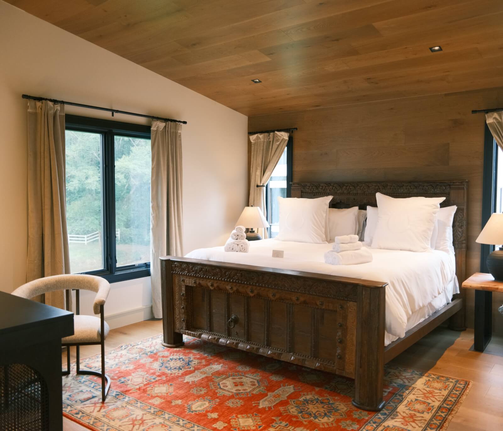 A cozy bedroom at a boutique farm near Hendersonville NC, with a large wooden bed, white bedding, two windows with beige curtains, a patterned rug, and a chair in the corner—perfect for a relaxing summer stay. Towels are placed on the bed.