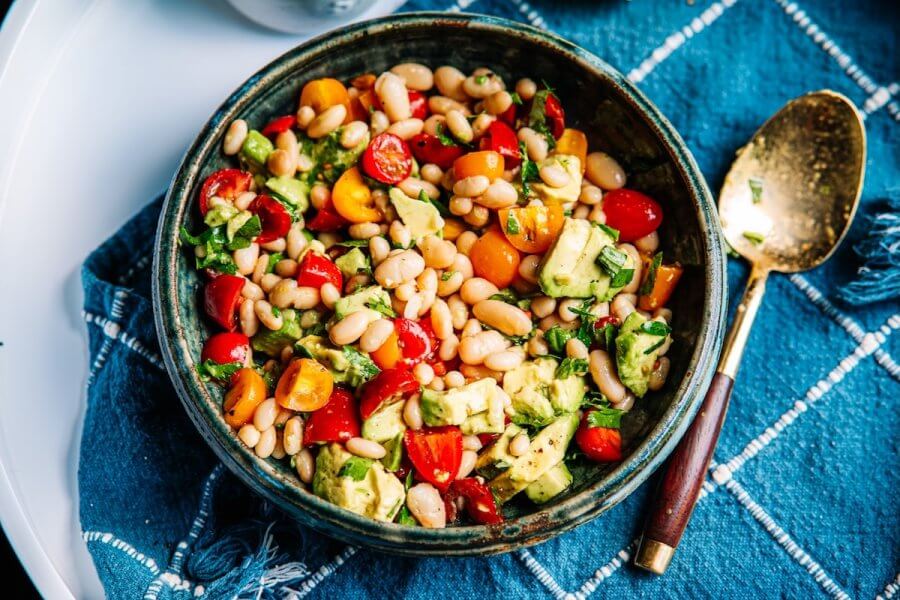 StyleBlueprint's 15 Most Popular Articles of 2025 - 12 A bowl of White Bean Salad with cherry tomatoes, avocado, and herbs sits on a blue cloth next to a gold spoon.