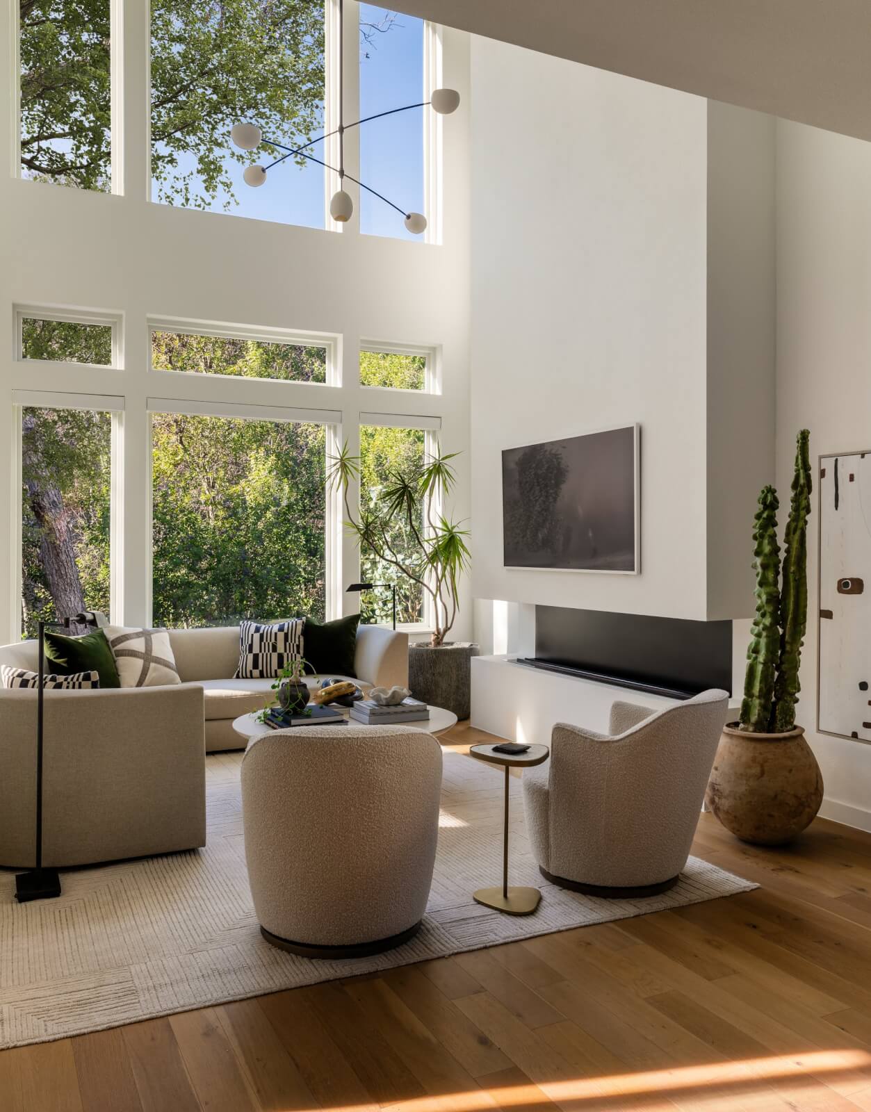 A modern living room with large windows, neutral furniture, sculptural design elements, indoor plants, a wall-mounted TV, and wood flooring. Natural light fills the space.