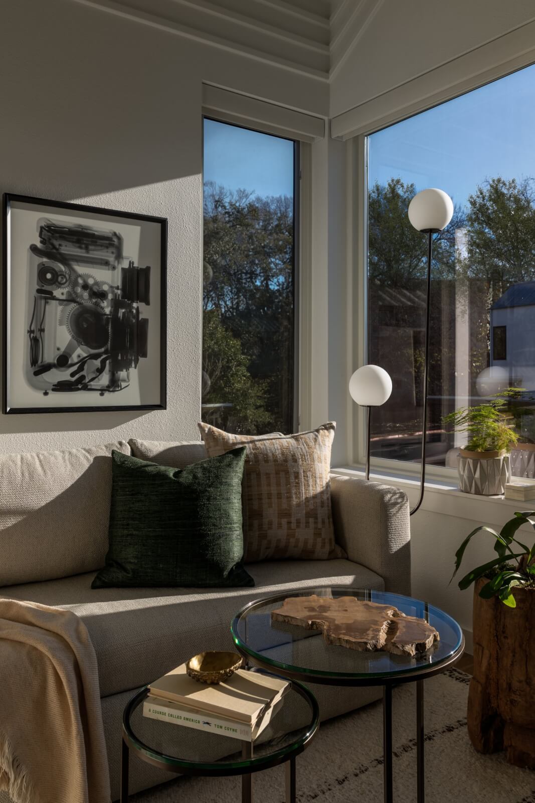 A modern living room corner with a light gray sofa, patterned pillows, glass coffee tables, and wall art exudes serene transformation; large windows reveal trees and buildings beyond.