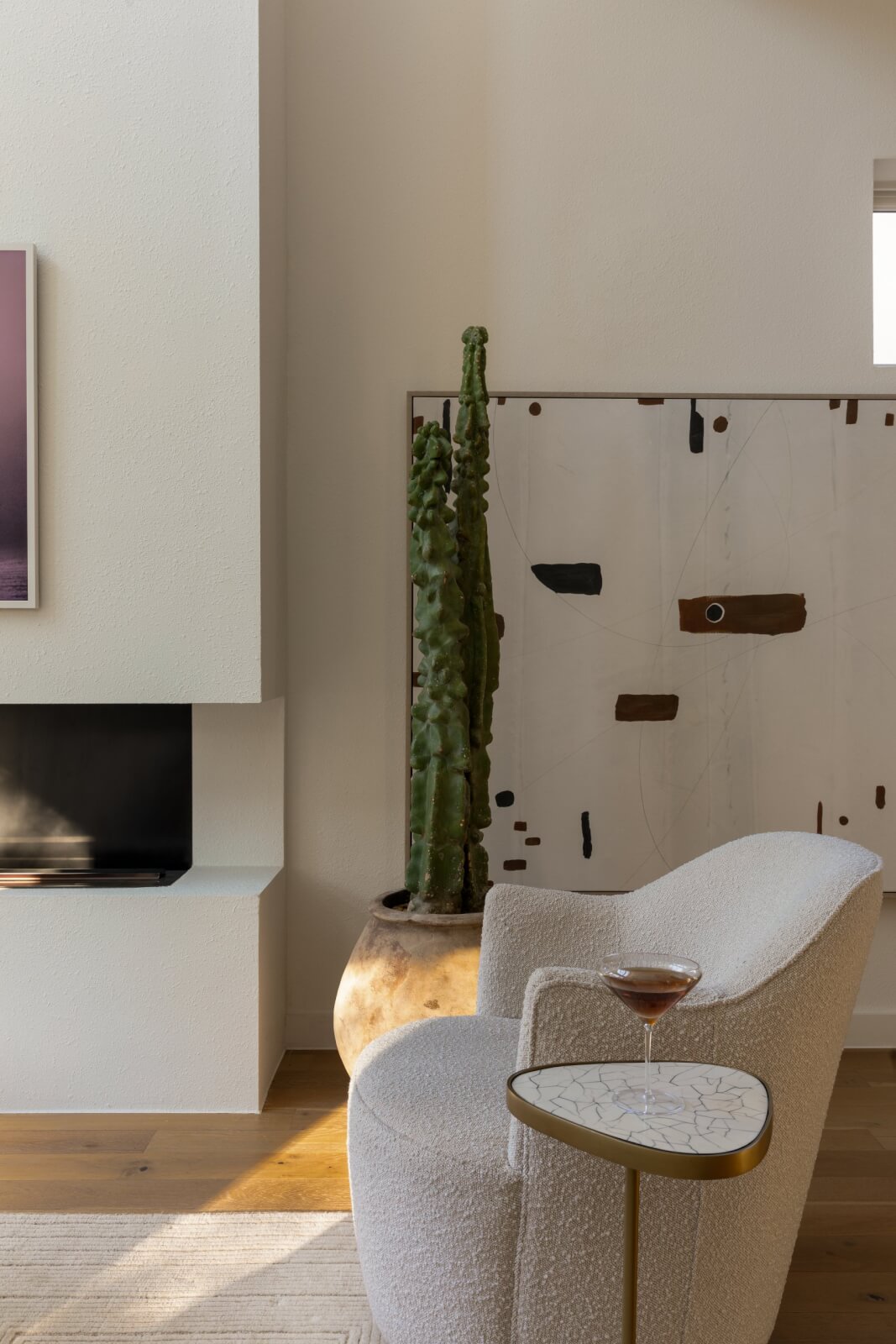 A modern living room featuring a white armchair, round side table with a drink, large cactus, and abstract wall art beside a built-in fireplace and TV reflects the serene sculptural design of this Austin home transformation.