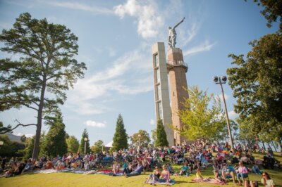 A crowd of people sits on blankets on a grassy lawn in front of a tall brick tower topped with a large statue, surrounded by trees under a clear blue sky.