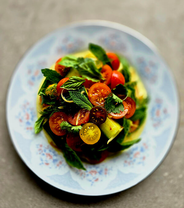 A plate of salad with sliced cherry tomatoes, cucumber ribbons, and fresh mint leaves arranged on a patterned white dish.