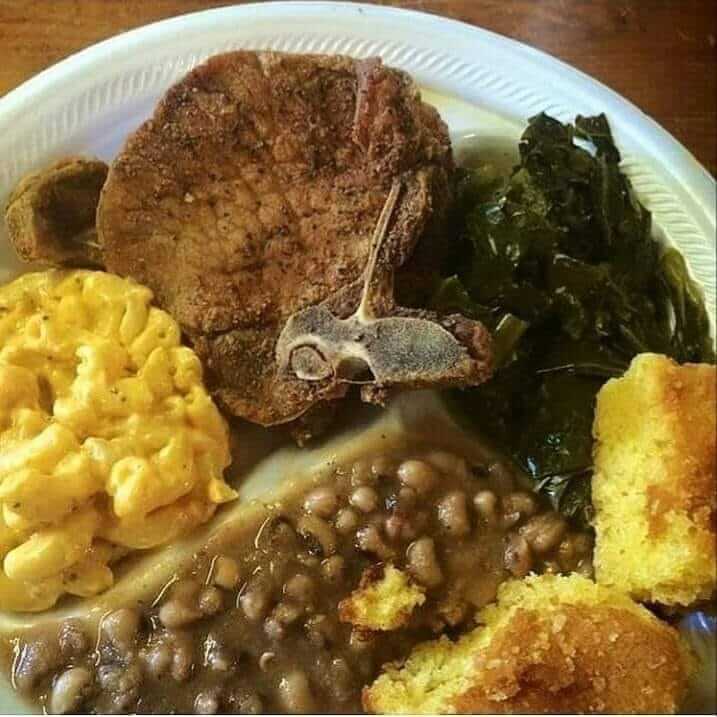 A plate with macaroni and cheese, pork chop, collard greens, cornbread, and black-eyed peas on a white disposable plate.